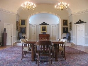 Image of the Philanthropic Hall looking towards the front of the room where there is a stage and desk. There is a large conference table in the foreground.